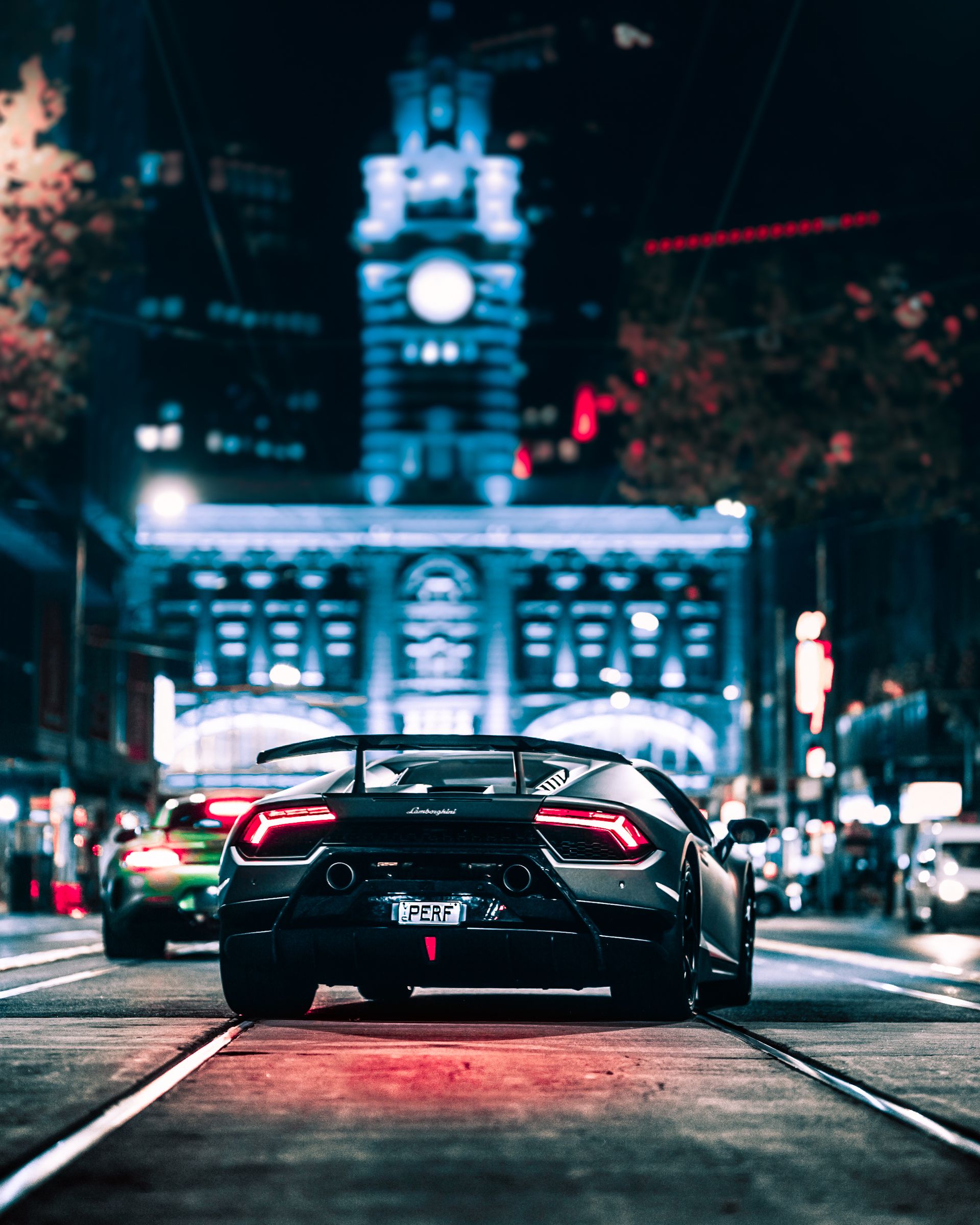 A super car parked on a city street at night, glowing under blue architectural lighting with red tail lights and gold ambient bokeh